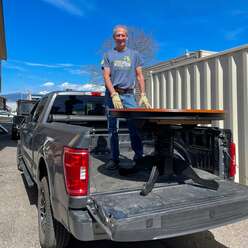 Man stands in flatbed of large black truck. He is smiling and standing behind a large wooden table.