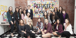 A group of people smiling for a picture in front of a sign that reads "Refugees Welcome."