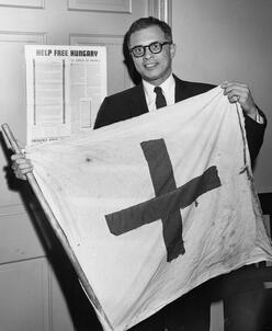 레오 체른은 "헝가리 해방을 도와주세요"라고 쓰인 푯말 앞에 서서 국기를 들고 사진을 찍기 위해 포즈를 취하고 있습니다.