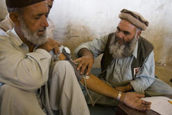 A doctor and another man, both Pakistani, sit on the floor, as the doctor takes the blood pressure of the other man.