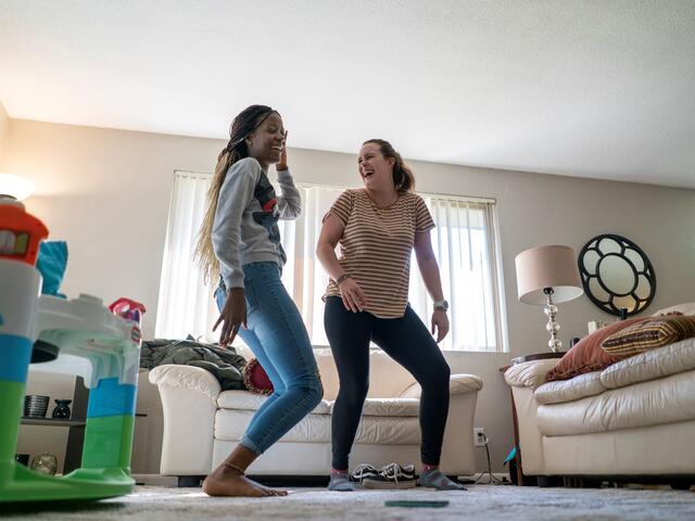 In the living room of Christelle’s apartment, Christelle teaches Charlee dance moves as the two laugh. There are children’s toys and two white couches also in the room. 