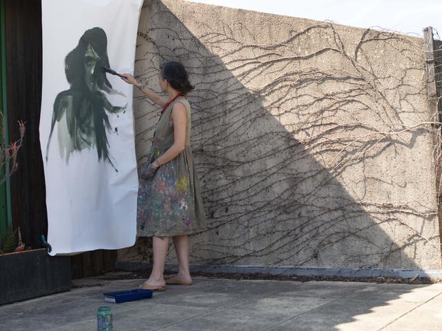 Diala Brisly, wearing a paint spattered smock dress, stads outside on a terrace painting a larger-rthan-life face on a sheet of paper hanging on the wall. 