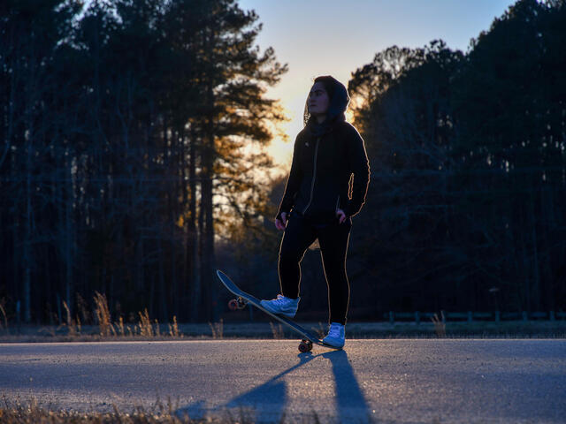 Belqisa stands on her skateboard in front of a forest sunset 
