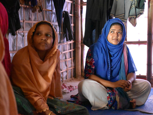 Two Rohingya women sit on mats, listening to an aid worker talk about monsoon preparedness in their refugee camp