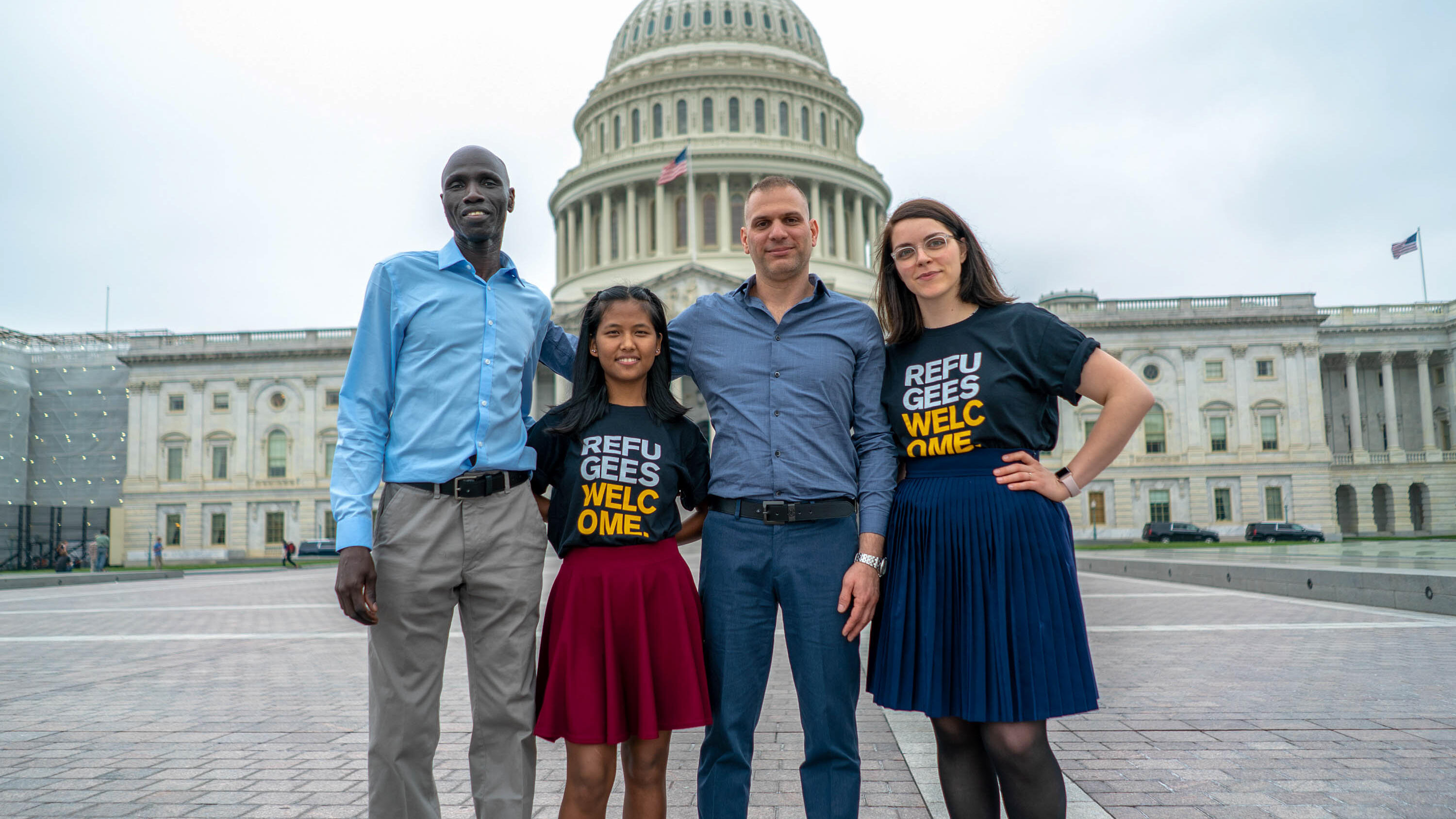 Four people pose for a picture in front of a capital building; two wear shorts with text reading "refugees welcome".