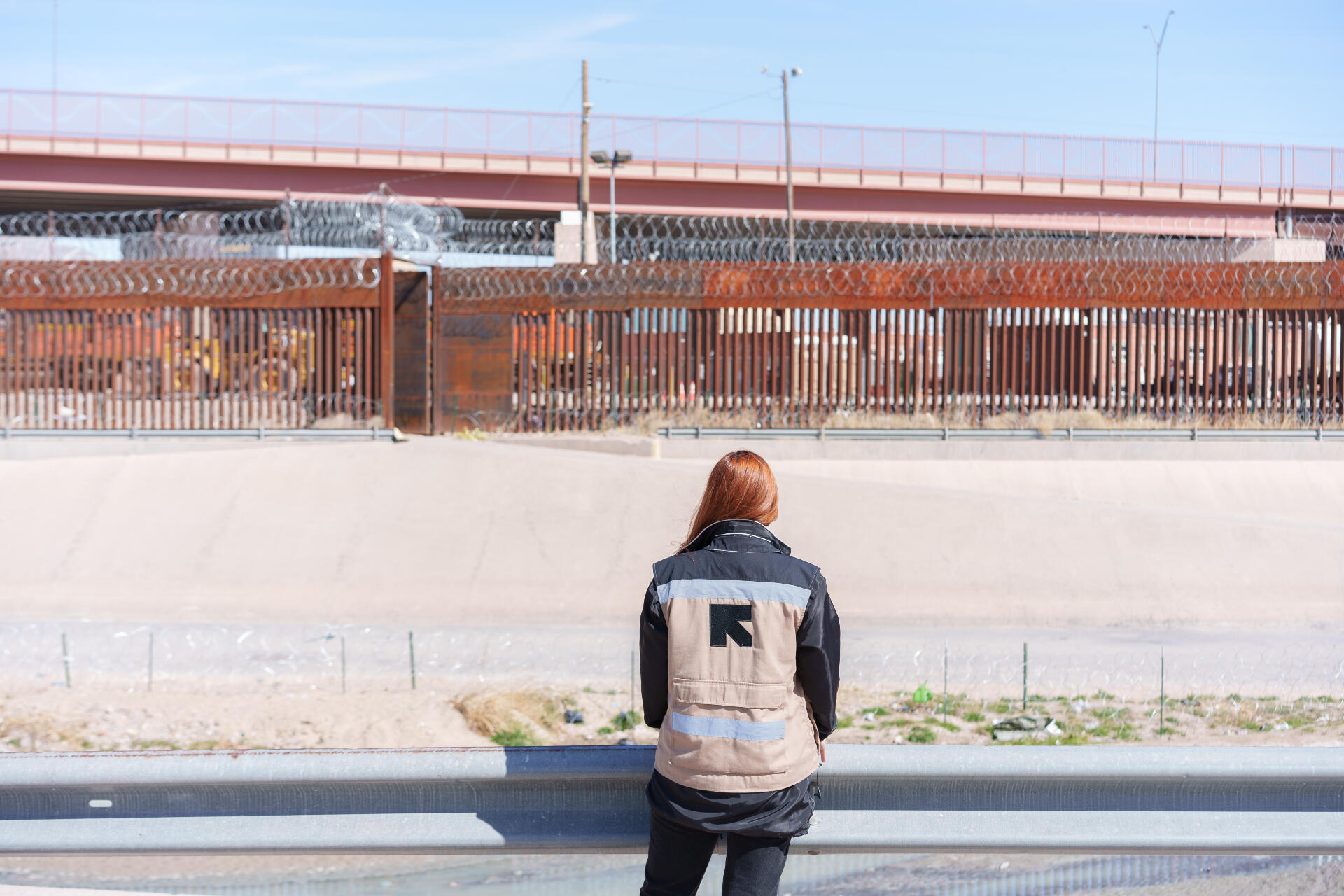 An IRC staff member faces away from the camera and towards the Mexico - U.S. border, while adorning a vest that has the IRC logo on the back.