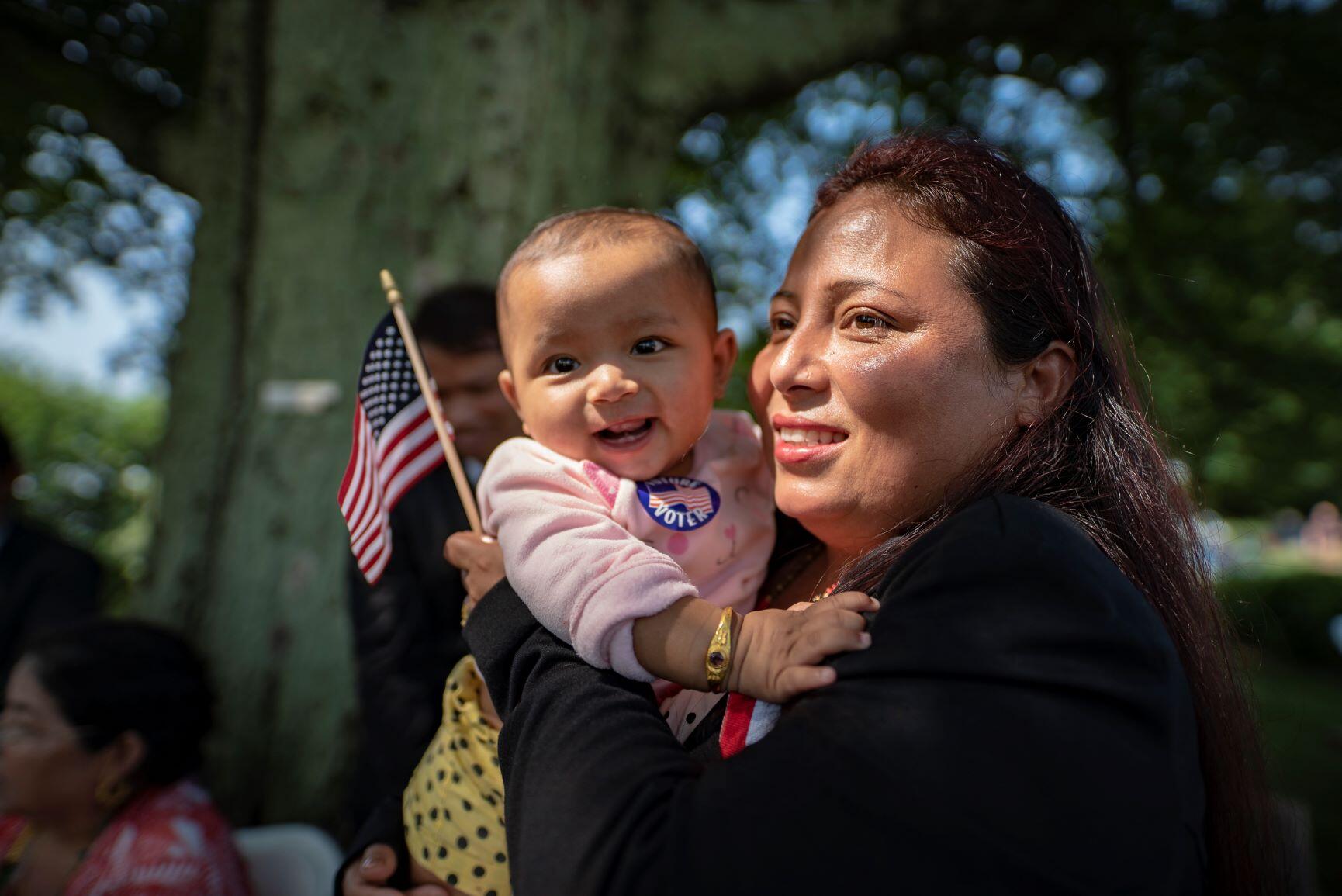 A newly naturalized citizen at a citizenship ceremony holds her baby and a small American flag. Both are smiling and the baby has a sticker on its shirt that says "Future Voter." 