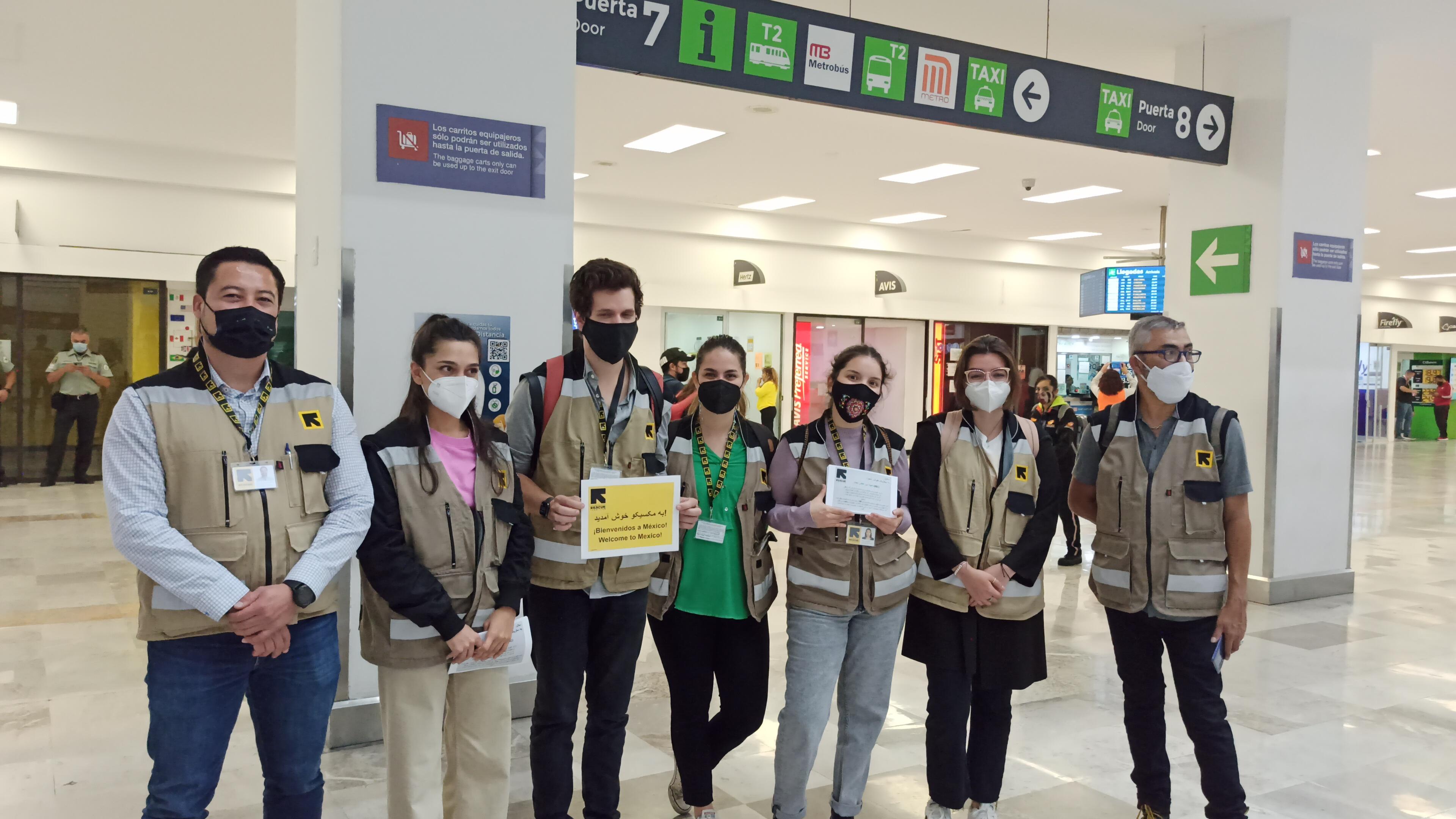 IRC staff members, all wearing masks and IRC-branded vests, stand in a line in an airport terminal. Some are holding welcome signs. 