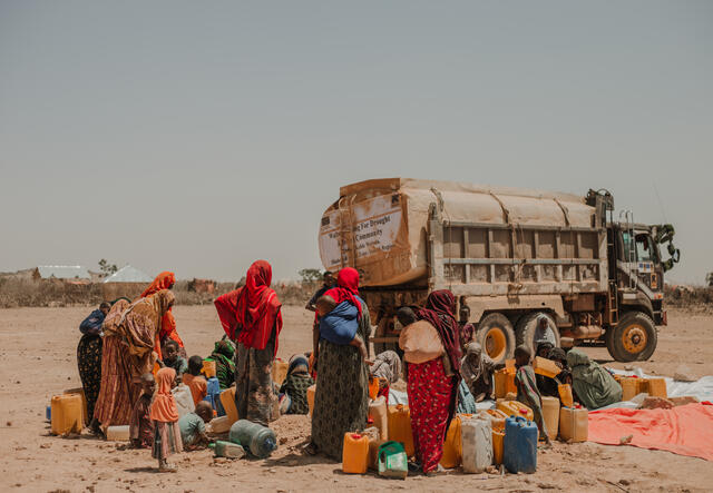 A group of women and children collect water from a truck amidst a barren and arid landscape.