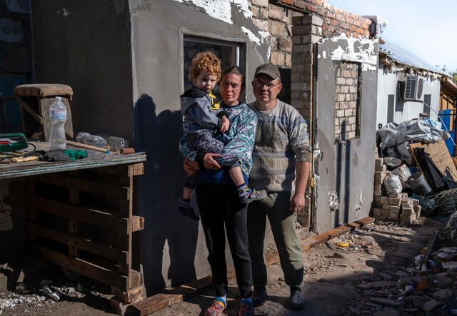 마리나가 아들을 안고 남편 옆에 서 있다. 배경에는 전쟁으로 파괴된 그들의 집이 보인다.