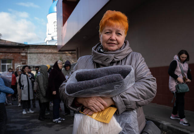 Lyubov faces the camera, holding a blanket she's been given by the IRC. Behind her, there's people are waiting in line.