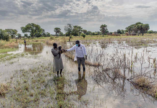 Abuk Deng holds her four year old daughter in her arms as they walk away from their flooded home while an IRC nutrition officer walks alongside them.