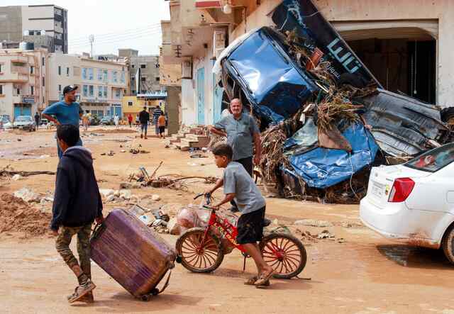 Two children are photographed in a flood-impacted region of Derna, Libya. One child pulls a suitcase while another pushes a bicycle.