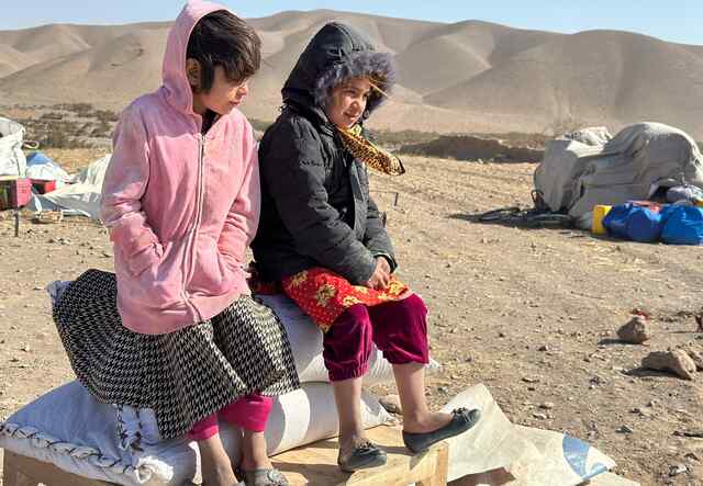 Two girls sit atop a small bench outside. In the background, Afghanistan's landscape is dotted with earthquake recovery supplies. 