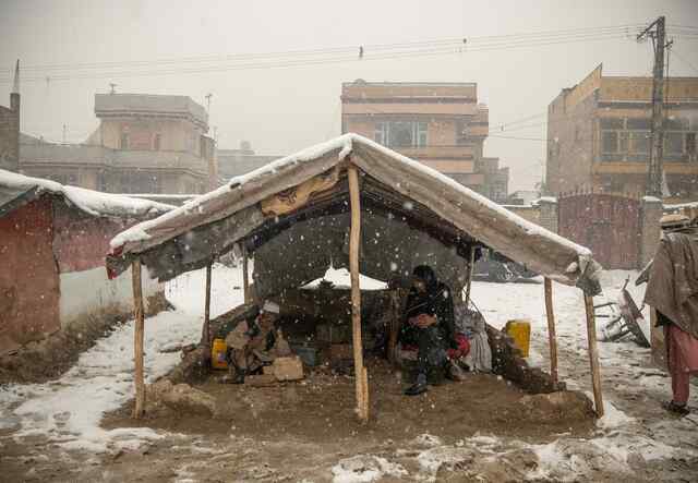 A man and a woman sit together in a small tent exposed to the element as heavy snow falls around them.