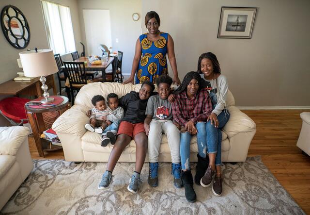 Jacqueline Uwumeremyi stands behind a couch where all of her children sit. There are 6 children, 4 boys and 2 girls.  