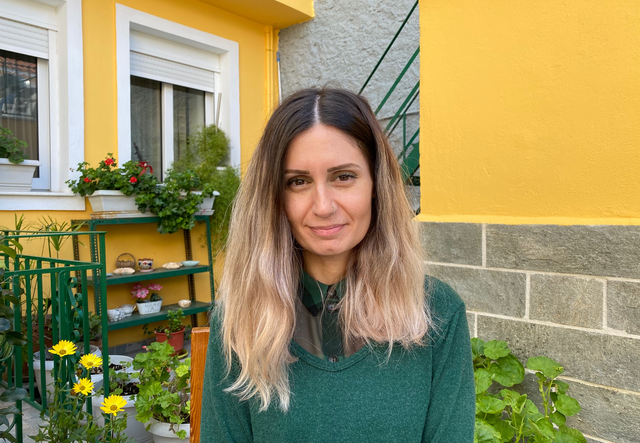 Kiki Michailidou, wearing a green sweater, sits outside a yellow house near a garden