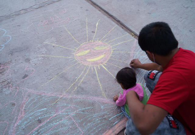 A father and young child bend over the ground drawing with chalk. They have already drawn a large sun. 