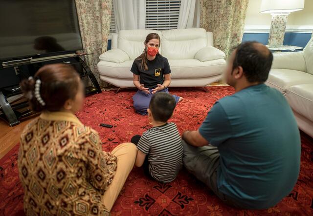 Jessica Carey, an IRC employment specialist, sits on the floor in a living room facing and talking to a family of three, parents and a young child.  