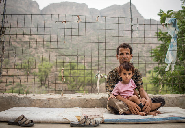 Against a view of a desert mountain, a man sits with his son on his lap. 