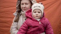 Iryna, a refugee from Ukraine, holding her daughter. They are standing in front of a tent and wearing all winter clothes. 
