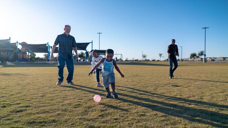 Dave, left, plays with Robert’s children, Sandra and Agape who call him Grandpa Dave. They are in a field and the children are kicking a ball. 
