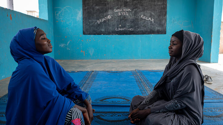 Falmata and Hauwa sit on a mat across from each other in an empty classroom practicing deep breathing mindfulness teachniques 