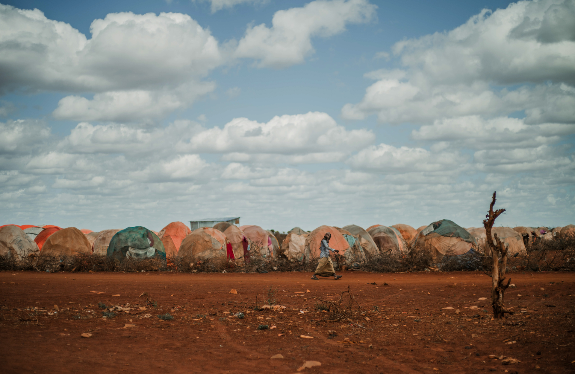 A wmen walks in front of a row of makeshift shelters in Somalia. The environment is barren and drought-stricken.