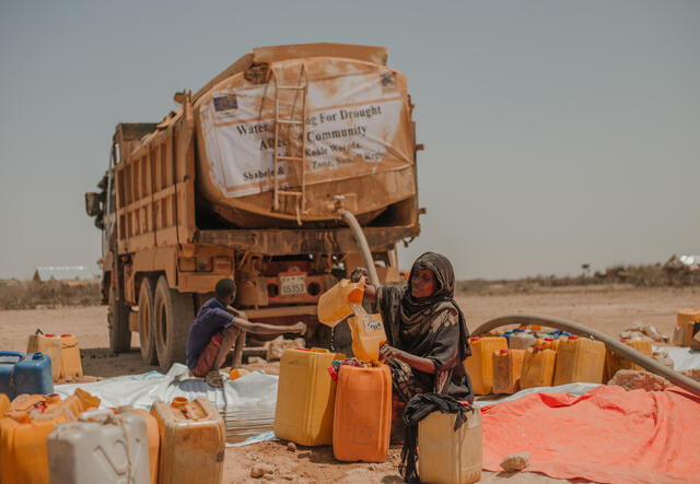 A woman sits and pours water from a truck into a large plastic container. Behind her is a desolate and dry landscape.