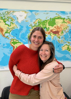 Two women stand in front of a world map smiling and hugging each other