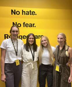 Four female Occupational Therapy students in business casual attire in front of a yellow wall with IRC badges on lanyards around their necks.