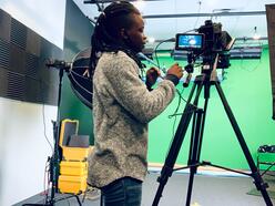 A sideprofile of Amen standing in front of a green background, focused on her video equipment