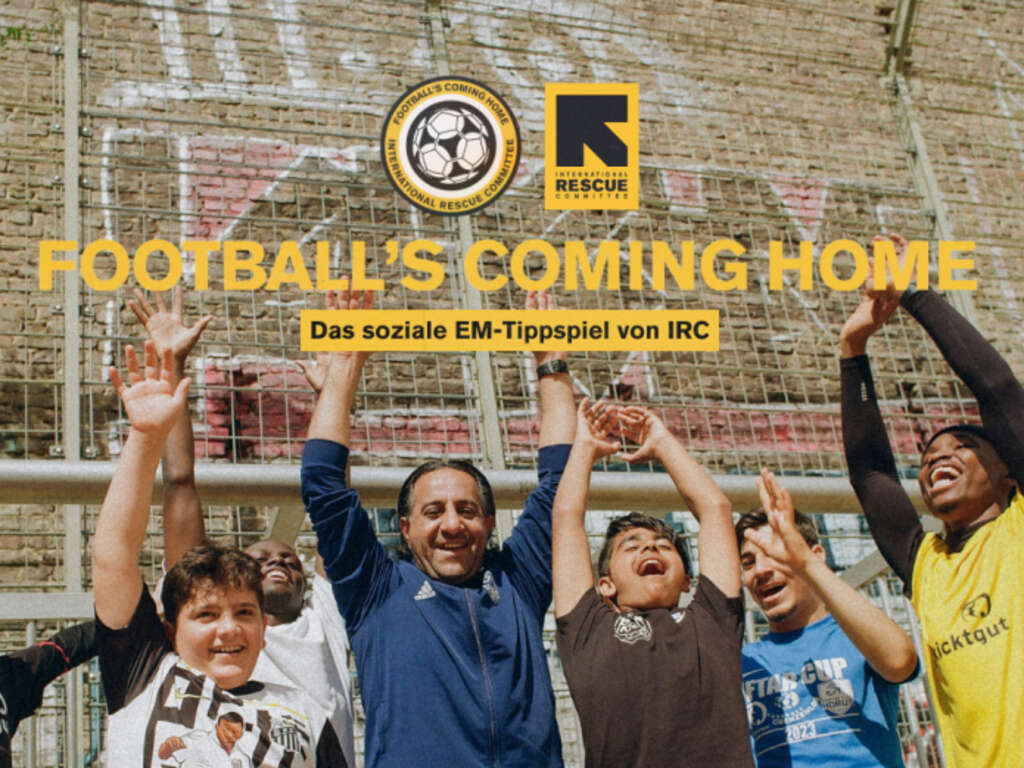 Eine Gruppe von Menschen jubelt unter dem Schriftzug „FOOTBALL'S COMING HOME“ mit Logos des „International Rescue Committee“ und der Aufschrift „Das soziale EM-Tippspiel von IRC“ im Hintergrund.