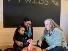 A young woman with a child on her lap sits across from an older woman. They are both smiling and looking down at the child who is playing with pink blocks.