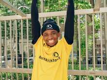 Ein junger Mann in einem gelben T-Shirt mit der Aufschrift 'buntkicktgut' und einer schwarzen Mütze hängt lächelnd an einer Stange.