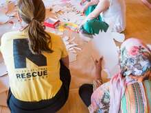 Rückansicht von drei Menschen, die an einem Kunstprojekt auf dem Boden arbeiten; eine Person trägt ein gelbes T-Shirt mit der Aufschrift 'Rescue Aid'.