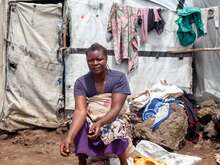 A woman sits outside, in front of a white makeshift shelter. A large pile of potatoes lie in front of her.