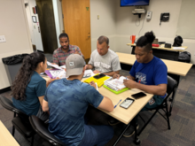 Adult students gather around a table with study materials, practicing their English language skills.