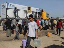 A man carrying water and walking away from the IRC water tank. Behind him there's a line of people waiting to do the same.