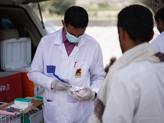 Pharmacist stands in front of open car boot writing on the box of medicine for a patient