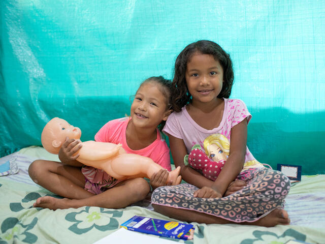 Two girls sit together smiling and holding dolls.