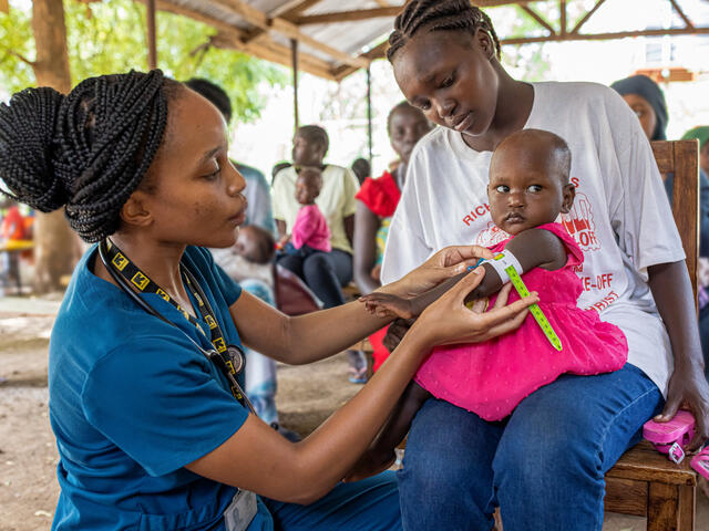 An IRC health worker screens a young girl for signs of malnutrition at an outdoor health clinic in Kenya. The child sits in her mothers lap while a malnutrition worker does the screening.