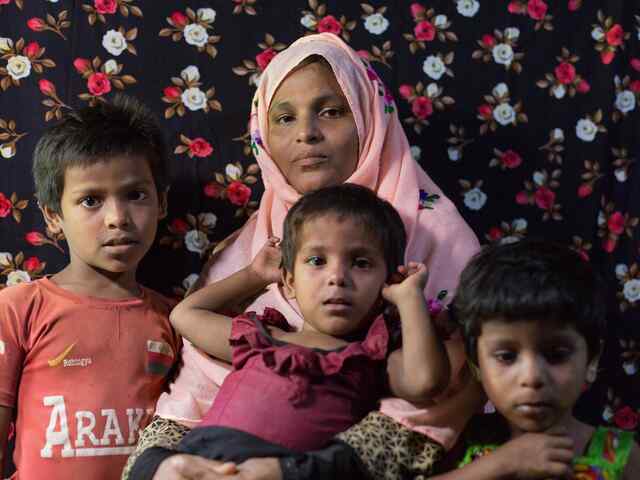 A mother, wearing a pink head scarf, holds one of her young children while two other children stand beside them.