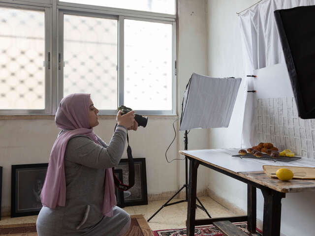 Hamida kneels, taking a photo of muffins on a table.