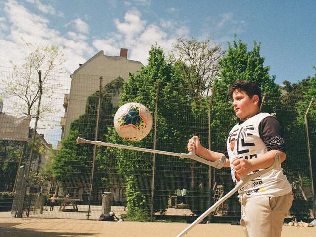 Ein Junge auf Krücken spielt Fußball auf einem sonnigen Spielplatz, konzentriert den Ball mit seinen Krücken jonglierend.