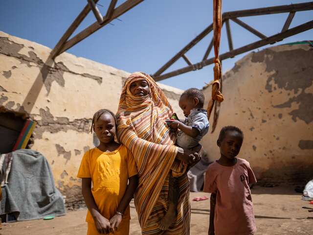 En mamma håller sitt yngsta barn i famnen. Två andra barn står vid sidan om henne i en skyddsbostad i Sudan.