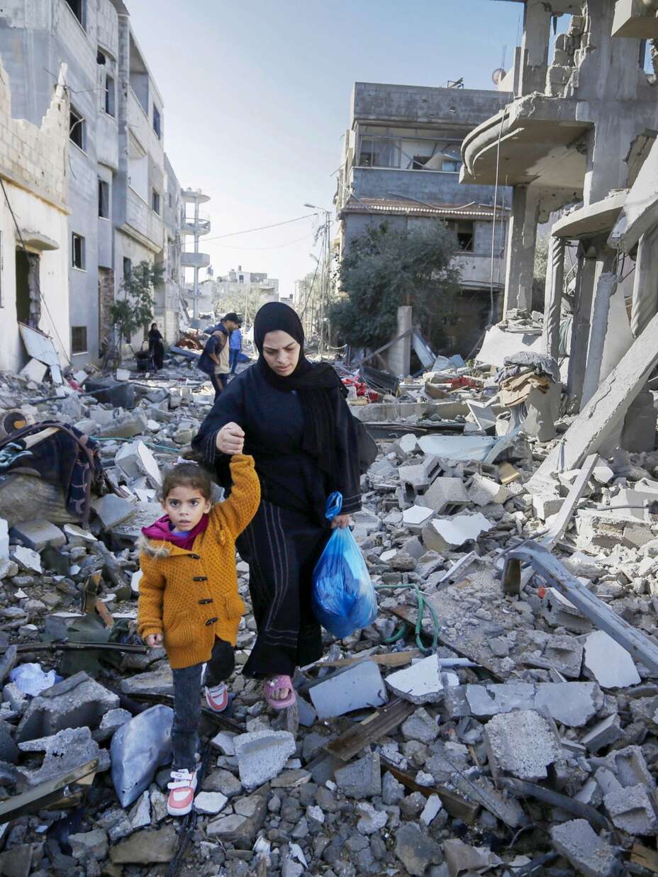 노란 재킷을 입은 어린 소녀와 그녀의 어머니는 전쟁으로 파괴된 가자 지구의 잔재를 걷고 있습니다.
