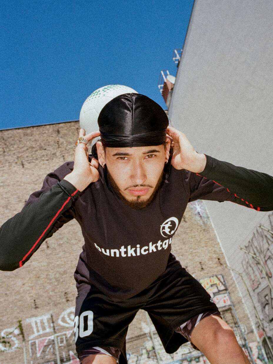Ein Mann in einem schwarzen „buntkicktgut“-Trikot balanciert einen Fußball auf seinem Kopf vor einer Wand mit Graffiti.