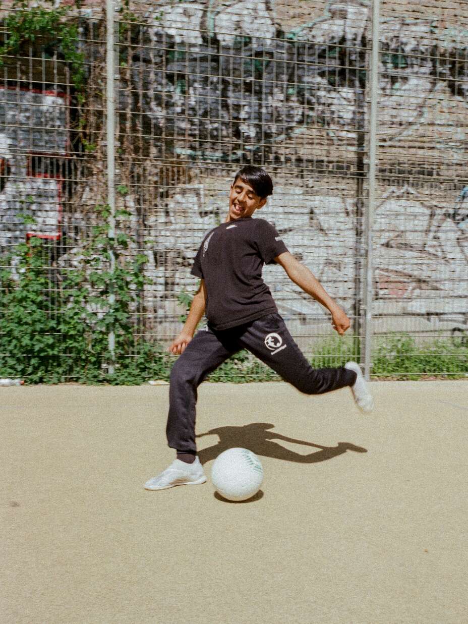 Ein junger Mann in einem schwarzen „buntkicktgut“-Trainingsanzug kickt einen Fußball auf einem Sportplatz vor einer Graffiti-bedeckten Wand.