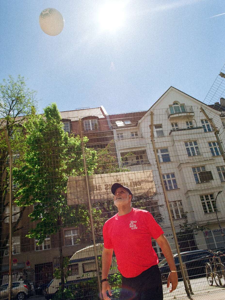 Ein Mann in einem roten T-Shirt und schwarzer Kappe schaut bei hellem Sonnenschein in die Luft, während ein Ball über ihm schwebt; im Hintergrund sind ein Zaun und mehrstöckige Gebäude zu sehen.